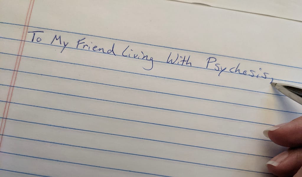 "To My Friend Living With Psychosis" is written in blue ink on lined paper with part of the author's pen and hand visible