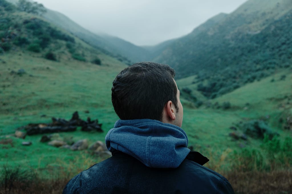 Man in a hoodie and jacket facing away from the camera looks at steep green hills with head slightly turned