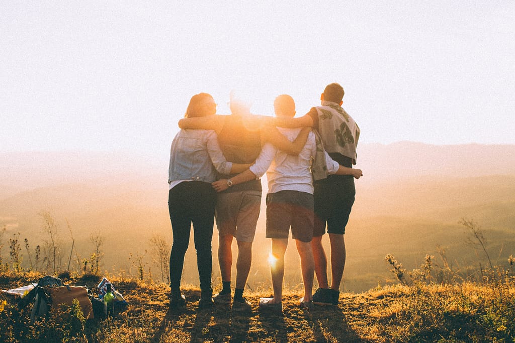 Four people stand at the top of a hill facing the sun with their arms around each others' shoulders.