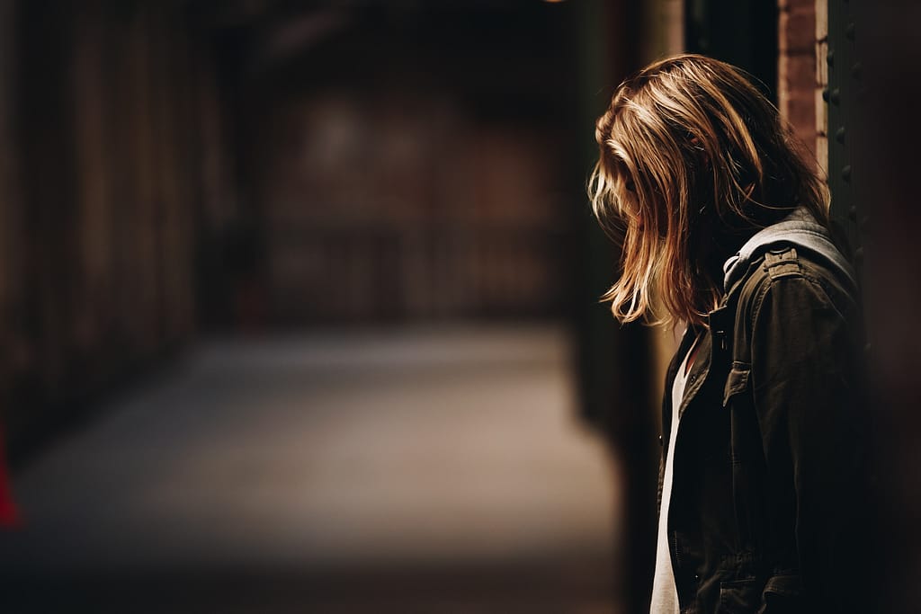 a girl with shoulder length blonde hair wearing a green jacket looks down at the ground with her hair covering her face in a shadowy hallway, possibly feeling the weight of invalidation.