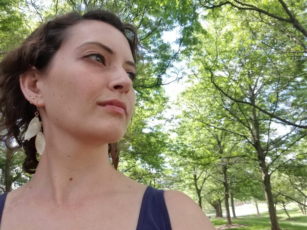 Katie, a woman with brown hair is looking to the side while wearing a blue tank top and gold earrings while outdoors under trees