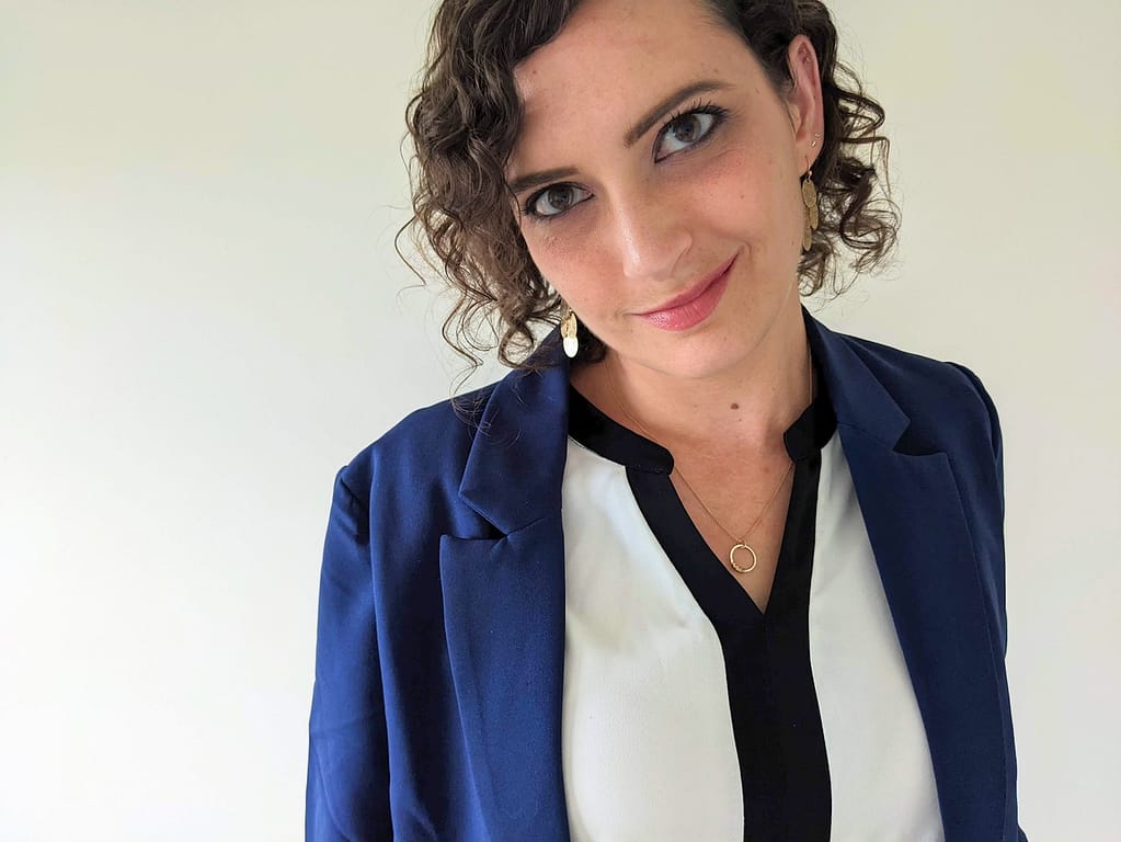 Katie, a woman with curly brown hair wearing a white and black blouse and blue blazer, looks into the camera as though she is looking at the professionals who should hire her.