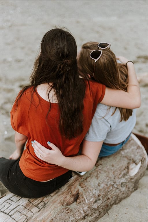 a woman with brown hair and a red tshirt and a woman with dark blonde hair, a grey tshirt, and sunglasses sit on a log with an arm around each other. This compassionate response is what speaking up about symptoms of mental illness should receive.