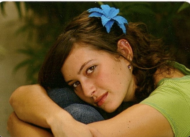 Katie, a young adult with brown curly hair and a green striped T shirt sits with her legs pulled to her chest with her arms wrapped around them and her cheek resting on her knees while wearing a fake blue flower in her hair.