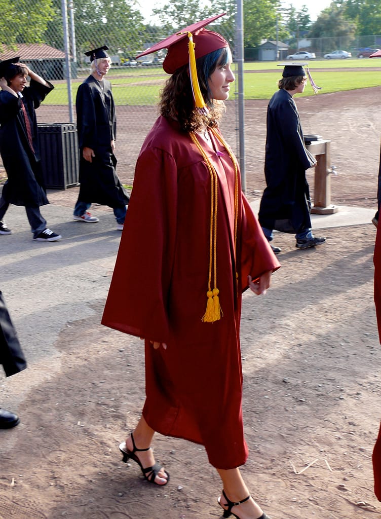 Girl with brown curly hair and blue streaks in her bangs wearing a dark read graduation gown and cap with yellow tassels walking into her high school graduation ceremony