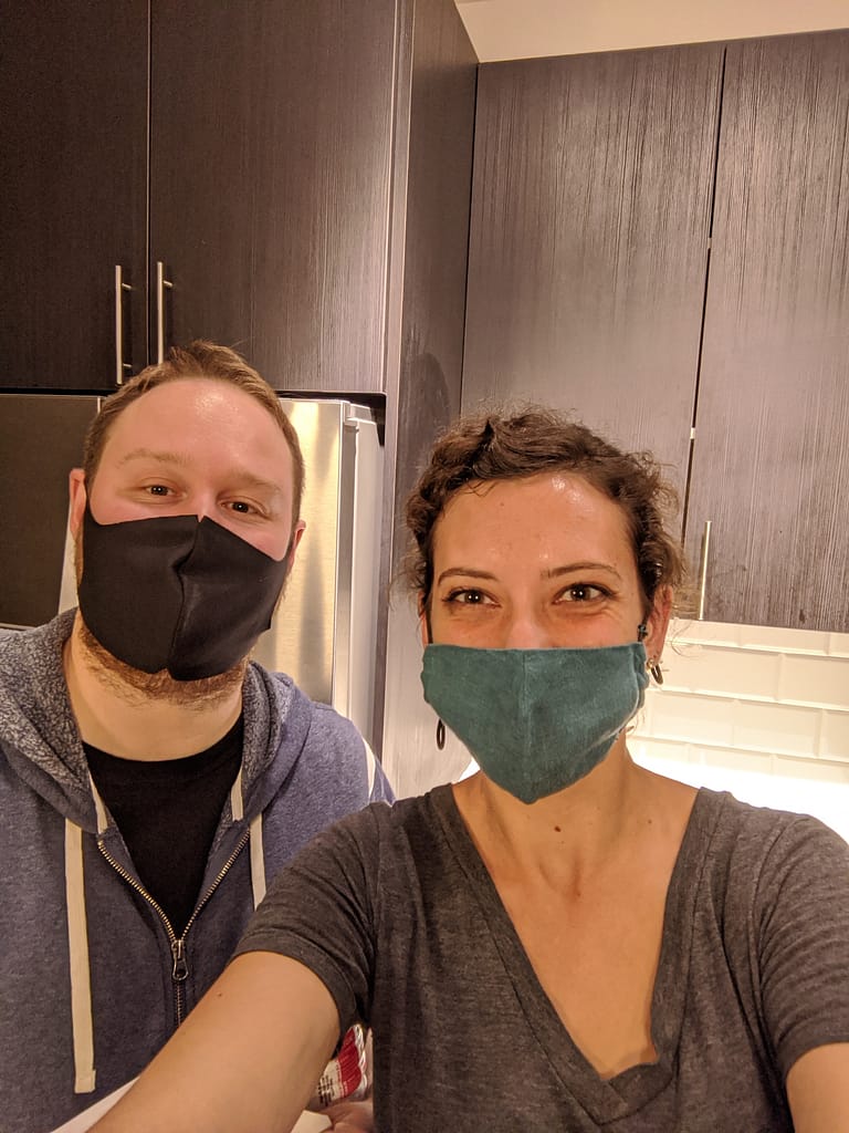 Katie and James stand in the kitchen of the empty apartment they just moved out of. James is wearing a black face mask and blue hoodie and Katie is in a teal mask and grey shirt.