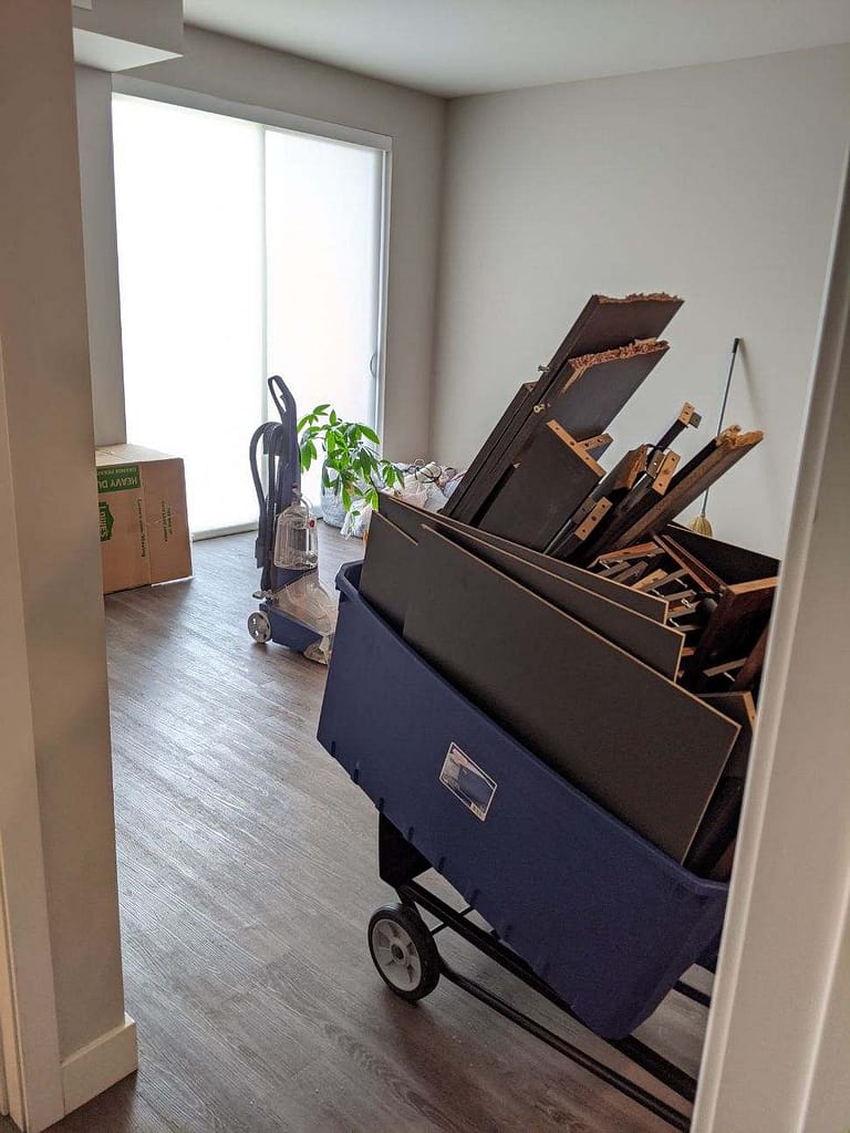 In the middle of moving day, the living room of Katie's apartment is empty other than a box, a carpet cleaner, a plant, and a blue bin of pieces of a bed