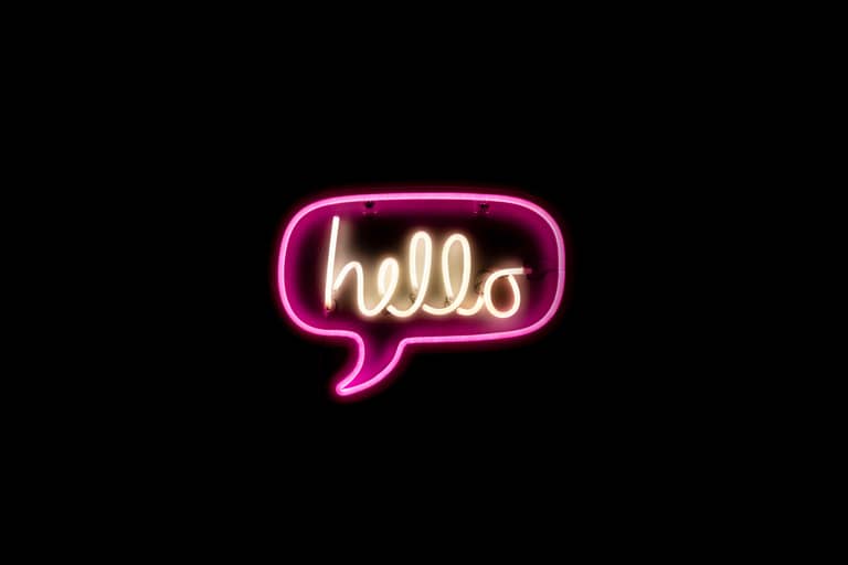 a neon sign reading "hello" in yellow in the center of a speech bubble with a pink outline is on a black background