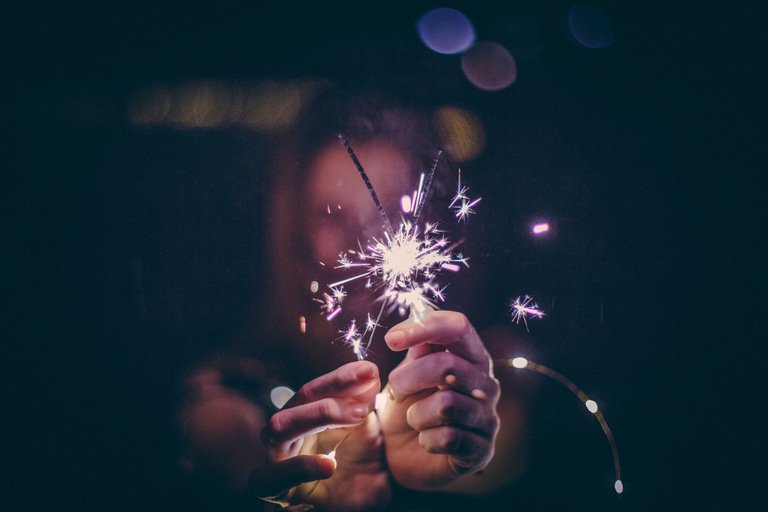 As another year ends, a woman holds a sparker firework in front of her with her face and body blurred other than her hands.