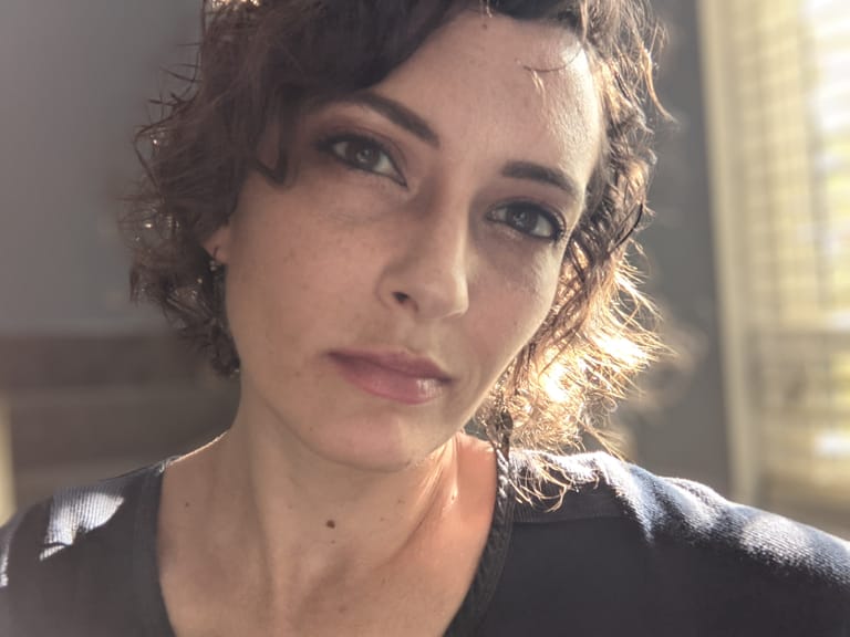 Katie, a woman with short brown curly hair and a dark blue shirt, looks into the camera with the sun lighting her face from the side as she thinks about life.