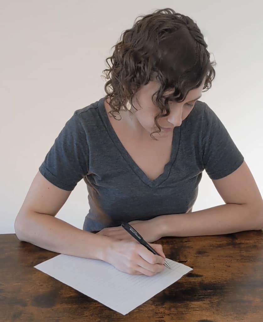Katie, a woman with brown curly hair wearing a grey shirt sits at a desk with a pen and paper writing Dear schizoaffective disorder