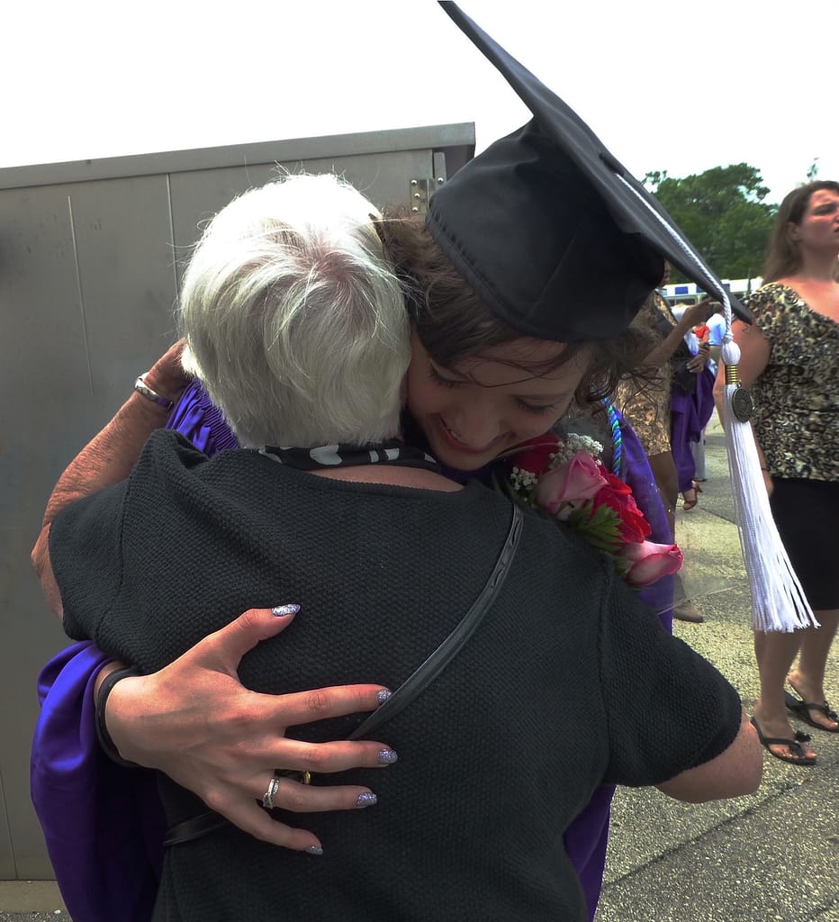 At Katie's college graduation, Katie, a woman with brown curly hair wearing a purple gown and black graduation cap, hugs Omi, a woman with short white hair wearing a black shirt